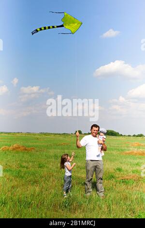 Glücklicher Mann, der Säugling und kleines Mädchen mit fliegendem Kite in der Nähe trägt, während er auf einem lebendigen Sommerrasen auf dem Feld steht Stockfoto