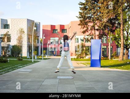 Teenager Schulmädchen in weißer Kleidung mit farbenfrohem Rucksack, die an sonnigen Tagen zur Schule geht und das Schulgebäude im Hintergrund hat Stockfoto