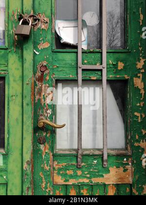 Nahaufnahme eines alten grünen Holztors mit abblätternder Farbe, Messinggriff, Metall-Sicherheitsgitter und einem Vorhängeschloss an einem verwitterten Glasfenster Stockfoto