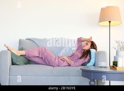 Junge Frauen in Schlafanzug genießen einen ruhigen Abend zu Hause entspannen Sie sich nach einem langen Tag auf der Couch und legen Sie auf ein graues Sofa neben dem Kaffeetisch mit Tasse o Stockfoto
