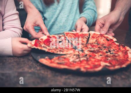 Schneiden Sie Eltern und Kinder zu, die köstliche Pizza mit Tomatensauce von einem großen Teller nehmen, während Sie gemeinsam ein Familienessen genießen Stockfoto