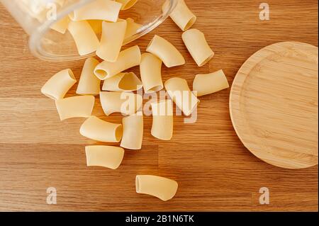 Verstreute trockene rohe große Makkaroni und Pasta aus einem großen Glasbecher mit einem Holzdeckel auf einer Holztischplatte, Draufsicht. Stockfoto