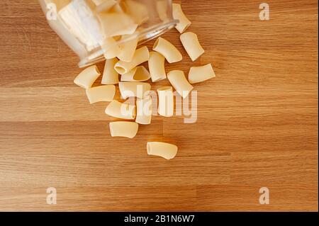 Verstreute trockene rohe große Makkaroni und Pasta aus einem großen Glasbecher mit einem Holzdeckel auf einer Holztischplatte, Draufsicht. Stockfoto