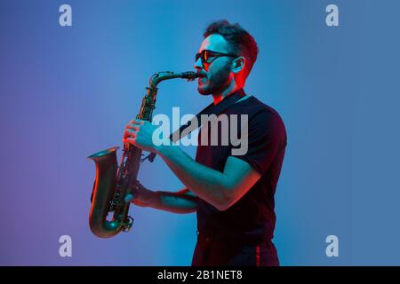 Junger kaukasischer Jazzmusiker, der das Saxofon auf einem gradienten blau-violetten Studiohintergrund in Neonlicht spielt. Konzept Musik, Hobby, Festival. Freudiger, attraktiver Kerl. Farbenfrohes Porträt des Künstlers. Stockfoto