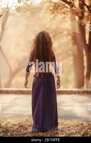 Ein raffiniertes schlankes Mädchen mit langen, lockigen Haaren in einem violetten Kleid mit Fußbodenlänge tanzt einen orientalischen Tanz vor dem Hintergrund des Herbstwaldes Stockfoto