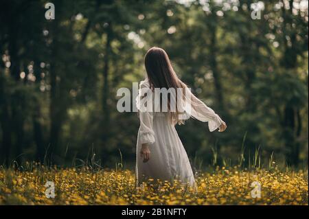 Ein Mädchen in einem weißen Baumwollkleid mit langen geraden Haaren genießt das Leben, dreht sich und tanzt in einem blühenden Feld gelber Blumen im warmen Sonnenuntergang Stockfoto