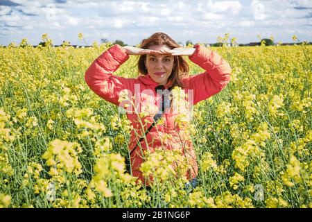 Schöne Frau in einem Rapsfeld, die ihre Augen von der hellen Sonne beschattete, mit beiden Händen das Sonnenlicht blockierte und dabei gerade in Richtung des blickte Stockfoto