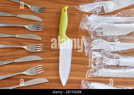 Großes Küchenmesser zwischen Einweg-Kunststoffsätzen und Edelstahl-Gabel- und Messerset, auf einer Nuss-Tischplatte. Konzept für Besteck aus Kunststoff im Vergleich zu reus Stockfoto