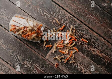 Mehrfarbige, spiralförmige, rohe Pasta zerbröckelten aus einem Glasbecher, um Produkte auf einer dunklen Holz-Tischplatte wiederverwendbar zu lagern Stockfoto