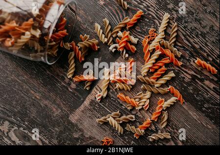 Mehrfarbige, spiralförmige, rohe Pasta zerbröckelten aus einem Glasbecher, um Produkte auf einer dunklen Holz-Tischplatte wiederverwendbar zu lagern Stockfoto