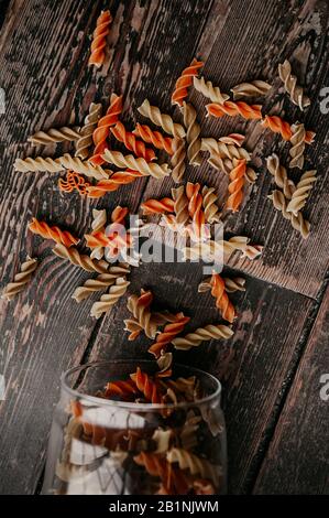 Mehrfarbige, spiralförmige, rohe Pasta zerbröckelten aus einem Glasbecher, um Produkte auf einer dunklen Holz-Tischplatte wiederverwendbar zu lagern Stockfoto