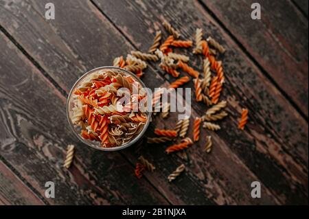 Die Option der intelligenten Aufbewahrung von Speisen in der Küche, ein Glasbecher zur Aufbewahrung von rohen Pasta und die verstreut gefärbten orangefarbenen Spiralpastas um sie herum Stockfoto