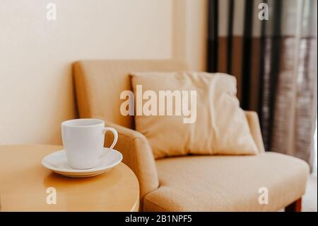Weiße Tasse auf einem Holzkaffeettisch vor dem Hintergrund eines beigefarbenen Sessels mit einem Kissen, eleganter Innenausstattung im Stil des Minimalismus, ein Ort für Stockfoto