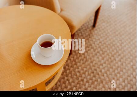 Ein weißer Becher, der halb voll Tee und mit einer weißen Untertasse auf einem runden Holzkaffeettisch ist, vor beigefarbenen Möbeln und einem braunen weichen Boden Stockfoto