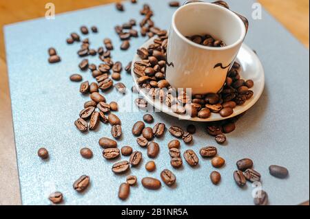 Der weiße Becher voller Kaffeebohnen steht auf einer weißen Platte, und die Körner sind verstreut Stockfoto