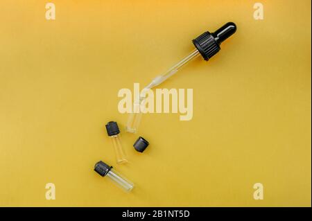 Drei kleine leere transparente Mixbecher mit schwarzem Deckel und Flüssigkeit tropft aus einer Pipette für die Kosmetologie oder Medizin auf einer einheitlichen, leuchtend gelben BA Stockfoto