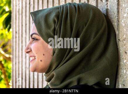 Ein Porträt des Profils einer attraktiven, lächelnden jungen muslimischen Frau, die vor neutralem Hintergrund einen dunkelgrünen Hijab trägt. Stockfoto