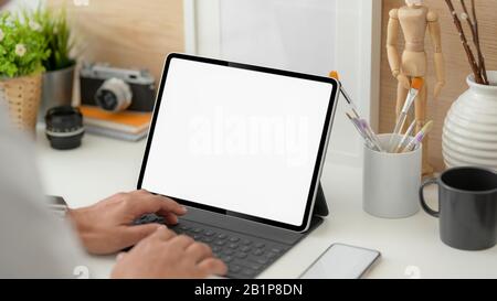 3/4-Aufnahme von jungen männlichen professionellen Designern, die auf einem verspotenen Tablet auf einem weißen Schreibtisch mit Stehrahmen, Kamera und Malwerkzeugen tippen Stockfoto