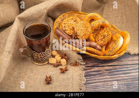 Plätzchen, Schokoladenkuchen, Bagels, Sternanis in einem Korbkorb und ein Glas Tee in einem Vintage-Cup-Halter vor dem Hintergrund rauer Homespun-Fabr Stockfoto