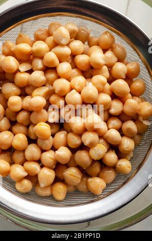 Oberirdische Aufnahme von Kichererbsen (Garbanzobohnen), die durch ein Stahlsieb in eine Glasschale auf einem weiß lackierten Holztisch ablaufen. Stockfoto