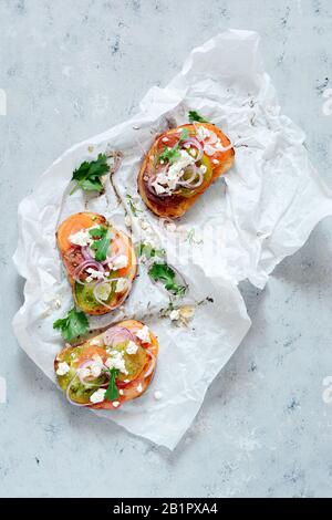 Italienische Bruschetta (Sandwich, Crostini, Tapas, Toast) mit frischen Tomaten, Ricotta, Kräutern, Zwiebeln, Olivenöl und Laub auf blauem Grund. Stockfoto