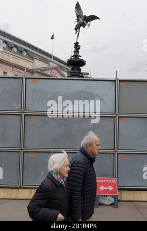 Ein älteres Paar geht an einer provisorischen Baustelle vorbei, die unter der teilweise verborgenen Statue des weltberühmten Wahrzeichen der viktorianischen Zeit in London, Eros im Piccadilly Circus, am 25. Februar 2020, in London, England, steht. Eros oder der Shaftesbury Memorial Fountain befindet sich an der Südostseite des Piccadilly Circus in London, Großbritannien. Nach dem zweiten Weltkrieg von seiner ursprünglichen Position im Zentrum versetzt, wurde es in den Jahren von 1892 bis 1893 zum Gedenken an die philanthropischen Werke von Lord Shaftesbury errichtet, der ein berühmter viktorianischer Politiker und Philanthrop war. Das Denkmal wird von Alfred Gilb überragt Stockfoto