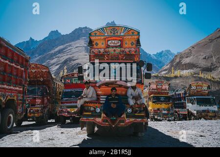 Sust, Pakistan - Oktober 2019: Drei Fahrer sitzen auf einem leuchtend orangefarbenen Truck, der mit bunten Mustern auf einem Bergparkplatz mit anderem TR dekoriert ist Stockfoto