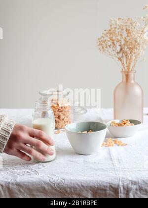 Hand der Frau, die eine Flasche Milch und eine Schüssel mit Cornflakes auf einem Frühstückstisch hält Stockfoto