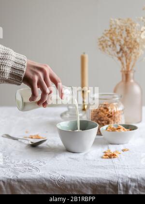 Die Hand der Frau gießt Milch in eine Schüssel mit Cornflakes. Helle Morgentischeinstellung mit getrockneten Blumen Stockfoto