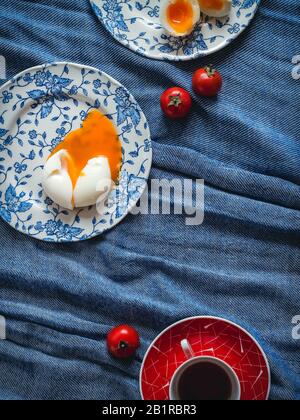 Weich gekochte Eier auf blau gemusterten Platten und rote Kaffeetasse auf blauem Leinen Stockfoto