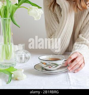 Detail einer jungen Frau, die mit weißen Tulpen in Vase und Tee am Tisch sitzt Stockfoto