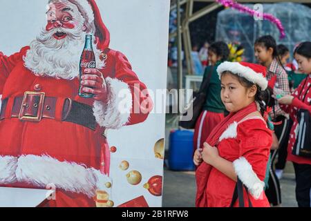 Während einer Weihnachtsfeier in Bangkok, Thailand, steht eine Schülerin des Weihnachtsmannes neben dem Plakat des Weihnachtsmannes, das Coca Cola wirbt Stockfoto