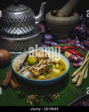 Schüssel aus traditionellem malaysischem soto oder Brühe mit Huhn, Nudeln und Gemüse, garniert mit Kalk umgeben von Zutaten Stockfoto