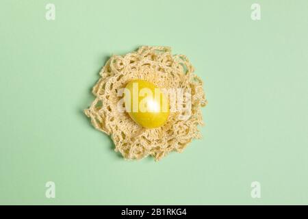 Ein gelb gestrichenes Osterei liegt auf gelber hausgemachter StrickServiette, gefaltet wie Nest auf trendigem Neo-Minze-Hintergrund. Minimalistischer Pastellbak aus Ostern Stockfoto