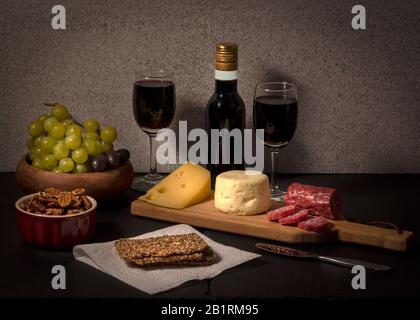 Brie und schweizer Käse auf schwarzem Steinkäsebrett, begleitet von Obst, Nüssen, Krackern und zwei Gläsern Rotwein, vor hellem Farbhintergrund Stockfoto