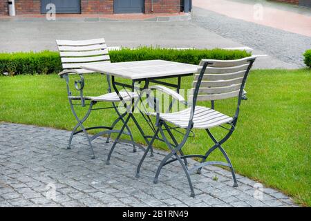 Holztisch mit eisernen Beinen und zwei Stühlen steht auf einem Kopfsteinpflaster in der Nähe des Rasens. Sommercafé. Stockfoto