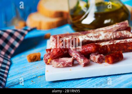 Einige Stücke Fuet und Chorizo, zwei verschiedene spanische Wurstwaren, auf einem weißen Teller auf einem blauen Holztisch, neben einigen Scheiben Brot an Stockfoto