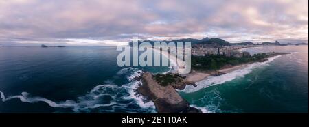 Wirbelndes Wellenmuster am Arpoador-Felsen in Rio de Janeiro, Brasilien, mit Ipanema-Strand und breiterem Stadtbild im Hintergrund vor einem bunten Himmel Stockfoto