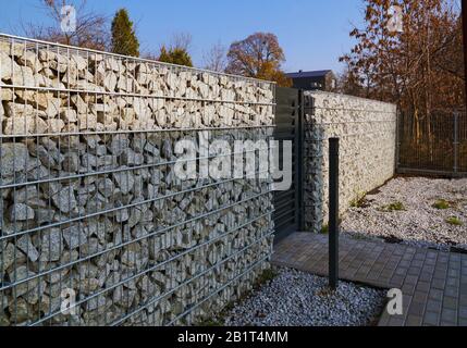 Zaun aus Gabionen. In der Wand, das Tor, vor dem Tor, ein Pfosten mit einem Knopf, um es zu öffnen. Stockfoto