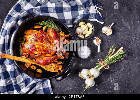 Vierzig Nelken gebratenes Hähnchen in einer schwarzen Backform auf einem Betontisch mit Knoblauch- und Thymianbund, horizontaler Blick von oben, flacher Lay, Kopie Stockfoto