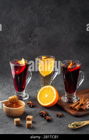 Drei große Gläser mit weißem und rotem Glühwein mit Orange, Zimtstäbchen, Sternanis, Honig in Holzschale, Rohrzuckerwürfel auf schwarzem Grund Stockfoto