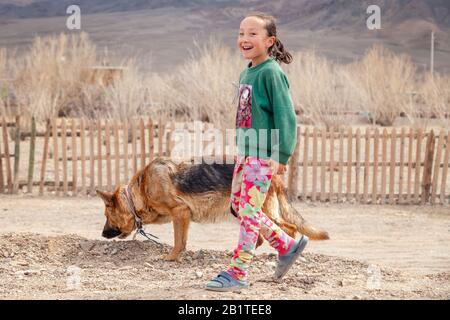 Mongolia Ulgii 2019-05-05 lächelndes mongolisches Mädchen in bunten Kleidern mit einem Hund kaukasischen Hirten vor dem Hintergrund von Zaun, Dorf, Bergen. Konzept Stockfoto