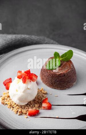 Weiße Platte mit Schokoladenkonditorkuchen mit Eis und Erdbeere auf dunklem Hintergrund, mit Minze und Sirup dekoriert, Blick von oben, vertikal Stockfoto
