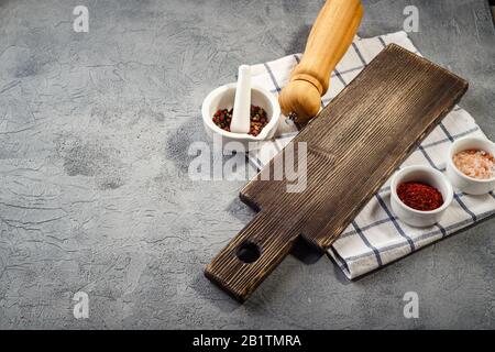 Holzschneidebrett und kleine Schalen mit Salz, Pfefferkörnern und Paprika, eine Gewürzmühle und ein Handtuch auf grauem Grund. Lebensmittelkonzept mit Platz f. Stockfoto