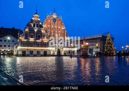 Blick auf das berühmte Haus der schwarzen Köpfe, das nachts beleuchtet wird, und auf den Weihnachtsbaum in Riga, Lettland. Blick auf den Rathausplatz und Rolands Statue Stockfoto