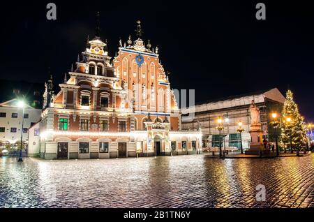 Blick auf das berühmte Haus der schwarzen Köpfe, das nachts beleuchtet wird, und auf den Weihnachtsbaum in Riga, Lettland. Blick auf den Rathausplatz und Rolands Statue Stockfoto