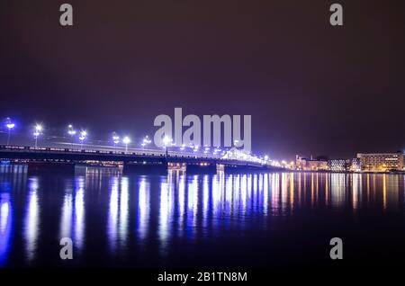 Blick auf die Steinbrücke in Riga in der Nacht, Lettland. Rigaer Steinbrücke am Abend. Beleuchtete Straße und Gebäude der Nationalbibliothek. Stockfoto