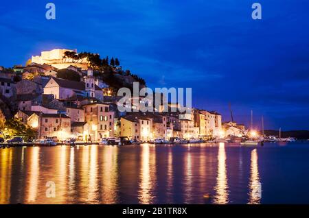 Beleuchtete Stadt mit Festung, alten Gebäuden, Meer und blauem Himmel. Hafen von Sibenik, Kroatien bei Nacht, Blick zur blauen Stunde auf die Festung St. Michael Stockfoto