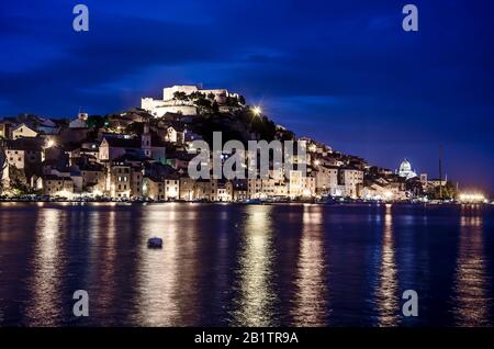 Beleuchtete Stadt mit Festung, alten Gebäuden, Meer und blauem Himmel. Hafen von Sibenik, Kroatien bei Nacht, Blick zur blauen Stunde auf die Festung St. Michael Stockfoto