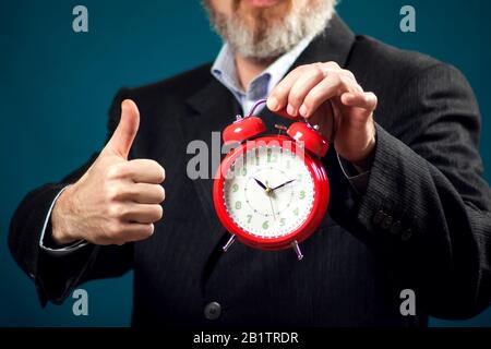 Bärtiger Geschäftsmann im Anzug, der roten Wecker hält und die Daumen nach oben zeigt. Zeitmanagement Konzept Stockfoto